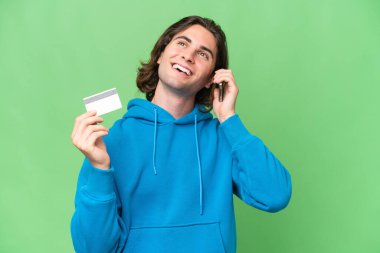 Genç yakışıklı adam yeşil renkli arka planda izole edilmiş cep telefonuyla sohbet ediyor ve elinde bir kredi kartı tutuyor.