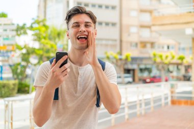 Dışarıda cep telefonu kullanan genç yakışıklı adam ağzı açık bağırıyor.