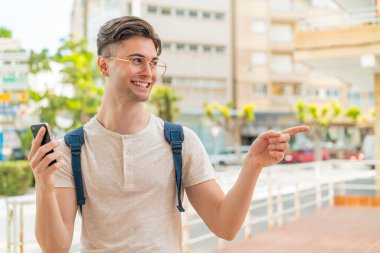 Dışarıda cep telefonu kullanan yakışıklı genç bir adam bir ürün sunmak için yan tarafı işaret ediyor.