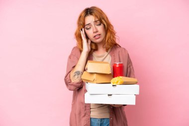 Fast food tutan beyaz bir kadın pembe arka planda baş ağrısıyla izole edilmiş.