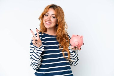 Beyaz arka planda izole edilmiş domuz kumbarası tutan beyaz bir kadın gülümsüyor ve zafer işareti gösteriyor.