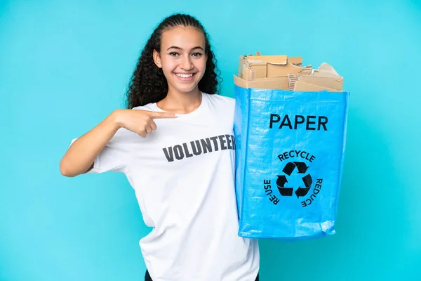 年轻的女人拿着一个装满了纸的回收袋 用蓝色的背景来回收 然后指着它 — 图库照片