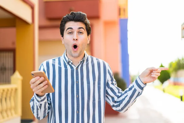 Mladý Pohledný Muž Pomocí Mobilního Telefonu Venku Překvapen Ukazuje Stranou — Stock fotografie