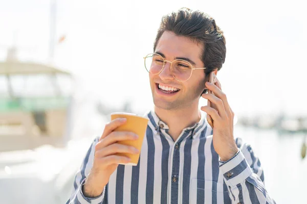 Mladý Pohledný Muž Pomocí Mobilního Telefonu Drží Kávu Veselým Výrazem — Stock fotografie