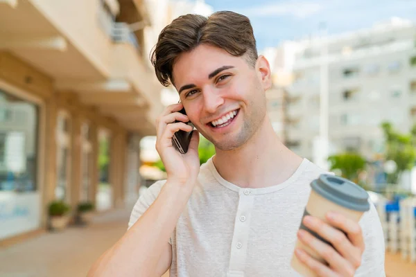 Mladý Pohledný Muž Pomocí Mobilního Telefonu Drží Kávu Veselým Výrazem — Stock fotografie