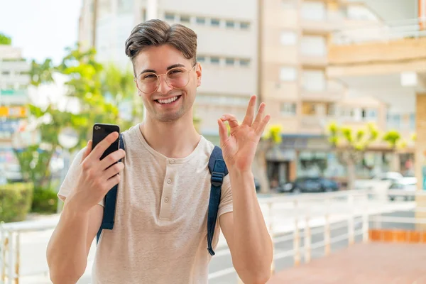 Mladý Pohledný Muž Pomocí Mobilního Telefonu Venku Ukazuje Znamení Prsty — Stock fotografie