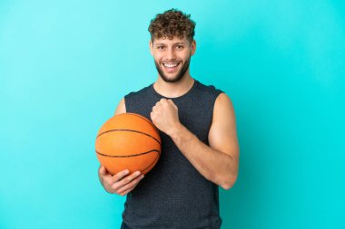 Basketbol oynayan yakışıklı genç adam mavi arka planda izole edilmiş bir şekilde zaferi kutluyor.