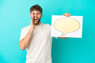 Genç beyaz adam mavi arka planda izole edilmiş. Elinde konuşma baloncuğu ikonu ve bağıran bir levha var.
