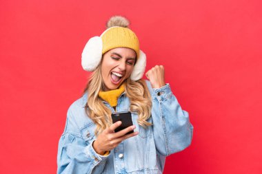 Kırmızı arka planda izole edilmiş kış kürkleri giyen genç ve güzel Uruguaylı kadın zafer pozisyonunda telefonla konuşuyor.