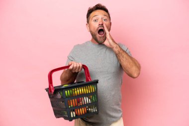 Elinde alışveriş sepeti olan orta yaşlı beyaz bir adam pembe arka planda şaşırmış ve şok olmuş bir yüz ifadesiyle izole edilmiş.
