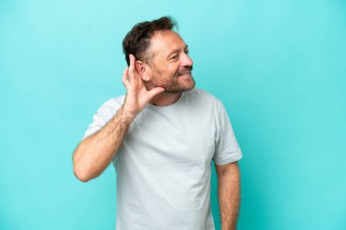 Middle age caucasian man isolated on blue background listening to something by putting hand on the ear