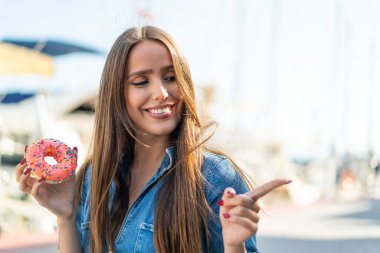 Açık havada donut tutan genç bir kadın bir ürün sunmak için kenarı işaret ediyor.