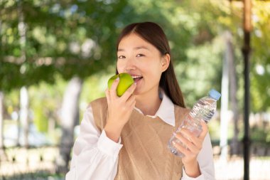 Güzel bir Çinli kadın bir şişe suyla dışarıda elma yiyor.