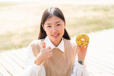 Genç Çinli bir kadın dışarıda çöreğini tutuyor. Başparmakları havada. Çünkü iyi bir şey oldu.