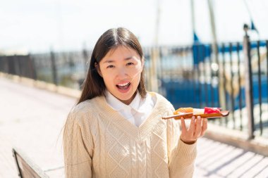 Genç Çinli bir kadın sürpriz ve şok olmuş bir yüz ifadesiyle açık havada saşimi tutuyor.