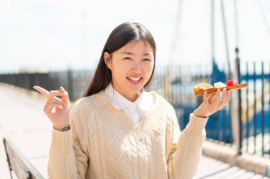 Genç Çinli kadın açık havada saşimi tutuyor. Şaşırmış ve parmağıyla işaret ediyor.