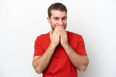 Beyaz arka planda izole edilmiş genç beyaz bir adam mutlu ve gülümseyen elleriyle ağzını kapatıyor.