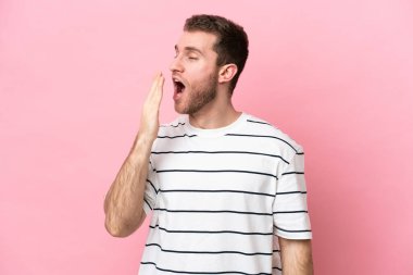 Pembe arka planda izole edilmiş genç beyaz adam esniyor ve ağzını eliyle açık tutuyor.