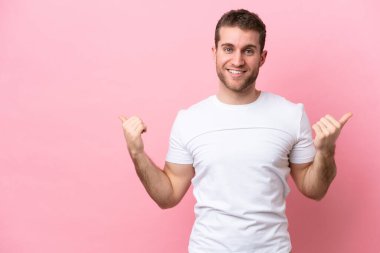 Pembe arka planda izole edilmiş genç beyaz bir adam. Baş parmakları havada ve gülümsüyor.