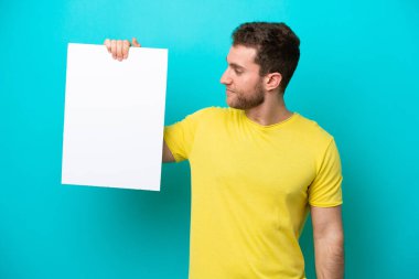 Kafkasyalı genç adam mavi arka planda izole edilmiş. Elinde boş bir levha var ve ona bakıyor.