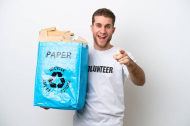 Beyaz arka planda izole edilmiş bir şekilde geri dönüşüm için elinde kağıt dolu bir torba tutan genç beyaz bir adam şaşırmış ve ön tarafı işaret ediyor.