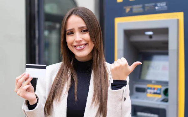 Young pretty woman holding a credit card at outdoors pointing to the side to present a product