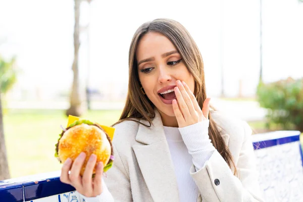 Młoda Ładna Kobieta Trzyma Hamburgera Świeżym Powietrzu Zaskoczeniem Zszokowanym Wyrazem — Zdjęcie stockowe