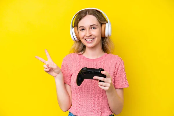 黄色い背景に孤立したビデオゲームコントローラーで遊ぶブロンド英国の若い女の子は笑顔と勝利のサインを示す — ストック写真