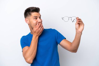 Young caucasian man holding glasses isolated on white background with surprise and shocked facial expression