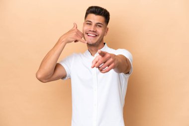 Beyazlı genç bir adam bej arka planda izole edilmiş telefon hareketleri yapıyor ve ön tarafı işaret ediyor.