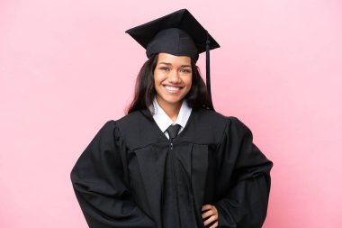 Kolombiyalı genç bir üniversite mezunu pembe arka planda izole edilmiş, kolları kalçasında ve gülümsüyor.