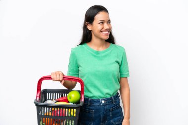 Beyaz arka planda izole edilmiş yiyecek dolu bir alışveriş sepeti tutan genç Kolombiyalı kadın.