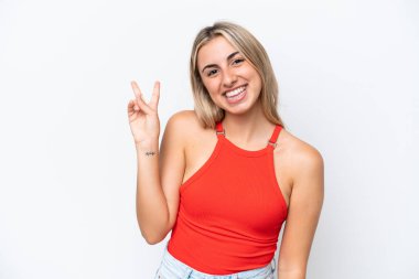 Beyaz arka planda izole edilmiş genç beyaz kadın gülümsüyor ve zafer işareti gösteriyor.