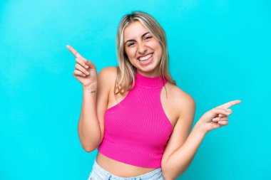 Mavi arka planda izole edilmiş genç beyaz kadın parmak uçlarını işaret ediyor ve mutlu.