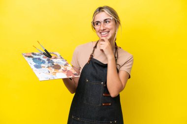 Young artist caucasian woman holding a palette isolated  on yellow background looking to the side and smiling