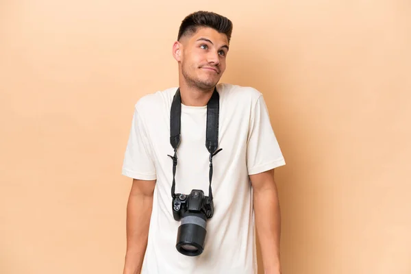年轻的摄影师高加索男子因米色背景而被隔离 提起肩膀时表示怀疑的姿态 — 图库照片