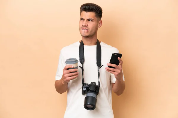 年轻的摄影师高加索人被隔离在米色背景下 一边拿着咖啡一边想着什么 一边拿着手机 — 图库照片