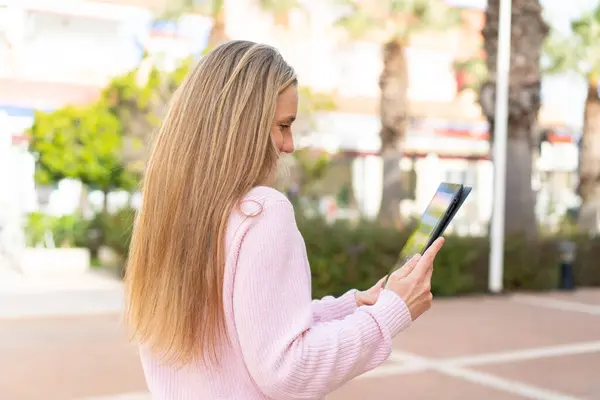 Young blonde woman holding a tablet at outdoors in back position