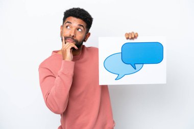 Genç Brezilyalı bir adam beyaz arka planda izole edilmiş. Elinde konuşma balonu ikonu olan bir levha var ve düşünüyor.