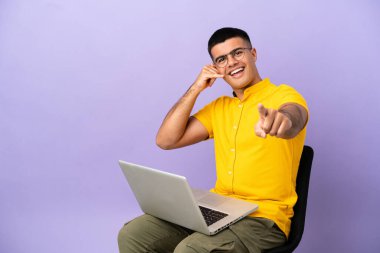 Genç adam dizüstü bilgisayarlı bir sandalyede oturuyor telefon hareketlerini yapıyor ve ön tarafı işaret ediyor.