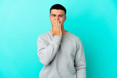 İzole edilmiş mavi arka planda genç yakışıklı adam ağzını eliyle kapatıyor.