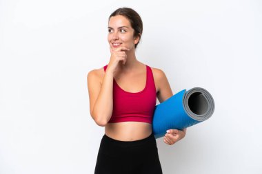 Beyaz arka planda izole edilmiş bir minderi tutarak yoga derslerine giden beyaz tenli genç bir kadın yan tarafa bakıyor ve gülümsüyor.