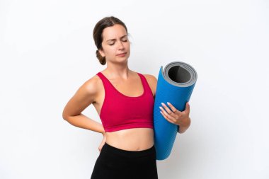 Beyaz arka planda bir paspas tutarken yoga derslerine giden beyaz, beyaz bir kadın çaba gösterdiği için sırt ağrısından muzdarip.