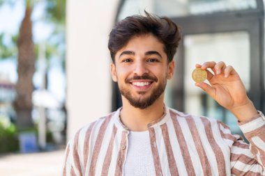 Yakışıklı bir Arap açık havada elinde mutlu bir ifadeyle Bitcoin tutuyor.