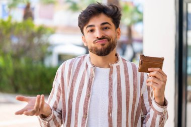 Yakışıklı bir Arap açık havada cüzdan tutuyor. Omuzlarını kaldırırken şüpheli hareketler yapıyor.
