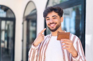 Dışarıdaki yakışıklı bir Arap, elinde mutlu bir ifadeyle cüzdan tutuyor.