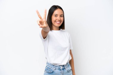 Beyaz arka planda izole edilmiş genç beyaz kadın gülümsüyor ve zafer işareti gösteriyor.