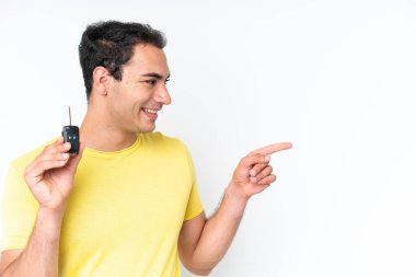 Beyaz arka planda izole edilmiş araba anahtarlarını tutan genç beyaz bir adam bir ürünü sunmak için yan tarafı işaret ediyor.