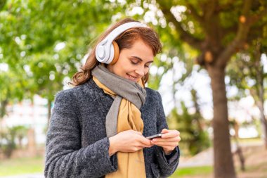 Açık havada müzik dinleyen esmer kadın cep telefonuyla mesaj yolluyor.