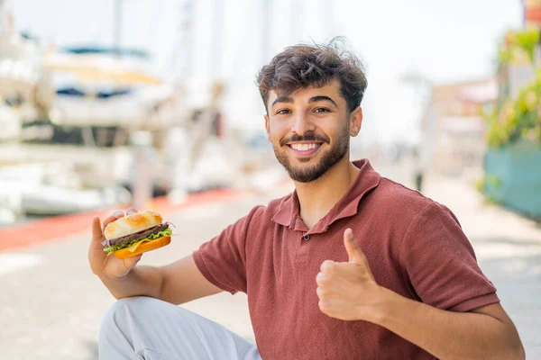良いことが起こったので 屋外でハンバーガーを抱えている若いアラビアのハンサムな男 — ストック写真